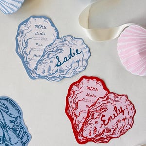 Peut inclure: Menus en forme d'huître bleues et rouges avec les noms "Sadie" et "Emily" en écriture cursive. Les menus présentent des entrées, des plats principaux et des desserts. Des découpes de coquillages et de homards bleus et roses sont également visibles.