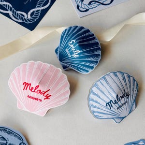 May include: Three seashell-shaped boxes in pink, blue, and white, each with a name in cursive. The pink box reads "Melody Ashworth" in red, while the blue box says "Emily Sweetness". The white box says "Melody Ashworth" in blue.