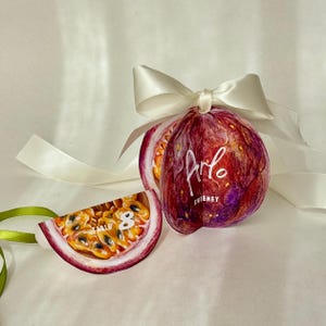 Könnte beinhalten: Ein dekoratives Passionsfrucht-Ornament mit einer weißen Schleife. Das Ornament ist tiefrot mit violetten Akzenten und dem Text "Arlo Sweeney". Ein Stück der Frucht ist ebenfalls zu sehen, das die Samen und das Fruchtfleisch zeigt.