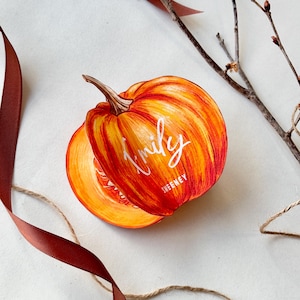 May include: A vibrant orange pumpkin illustration featuring a brown stem and visible ribbing. The name Emily is written in white cursive script across the centre, with the name SWEENEY printed in smaller white capital letters underneath.