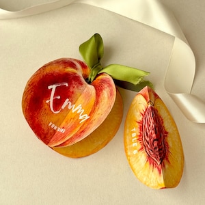 Può includere: Segnaposto a forma di pesca con il nome "Emma" e "For Wine" in scrittura bianca. Un segnaposto è tagliato per rivelare l'interno della pesca, con un nastro verde e un nastro bianco sullo sfondo. Il secondo segnaposto ha il testo "Table 6".