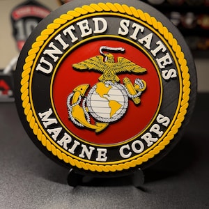 May include: A round black emblem with the words "UNITED STATES MARINE CORPS" in white lettering. The center features a red background with a gold eagle, globe, and anchor emblem. A yellow rope-like border surrounds the emblem.