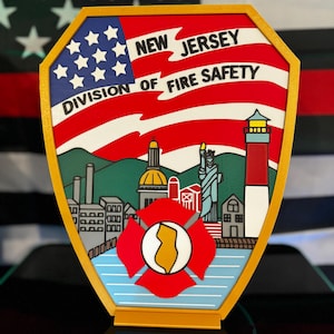 May include: A gold-framed emblem featuring the words "New Jersey Division of Fire Safety" with a flag design. The emblem includes illustrations of a cityscape, a lighthouse, and the Statue of Liberty. A red and yellow fire department emblem is in the center.
