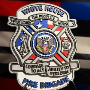 May include: A multi-layered, shield-shaped emblem featuring the text WHITE HOUSE, THE PEOPLE'S HOUSE, PROTECTING, COURAGE TO ACT, ABILITY TO PERFORM, and FIRE BRIGADE. The center displays a bald eagle seal, with icons of a ladder and fire hydrant.