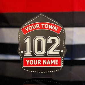May include: A black shield-shaped sign with white stitching detail. It features a red banner at the top with the text YOUR TOWN, the large white number 102 in the center, and a red banner at the bottom with the text YOUR NAME.