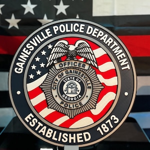 May include: A circular Gainesville Police Department emblem featuring a central badge with an eagle, the text City of Gainesville State of Georgia Police, and the words Gainesville Police Department Established 1873 on a red, white, and blue background.