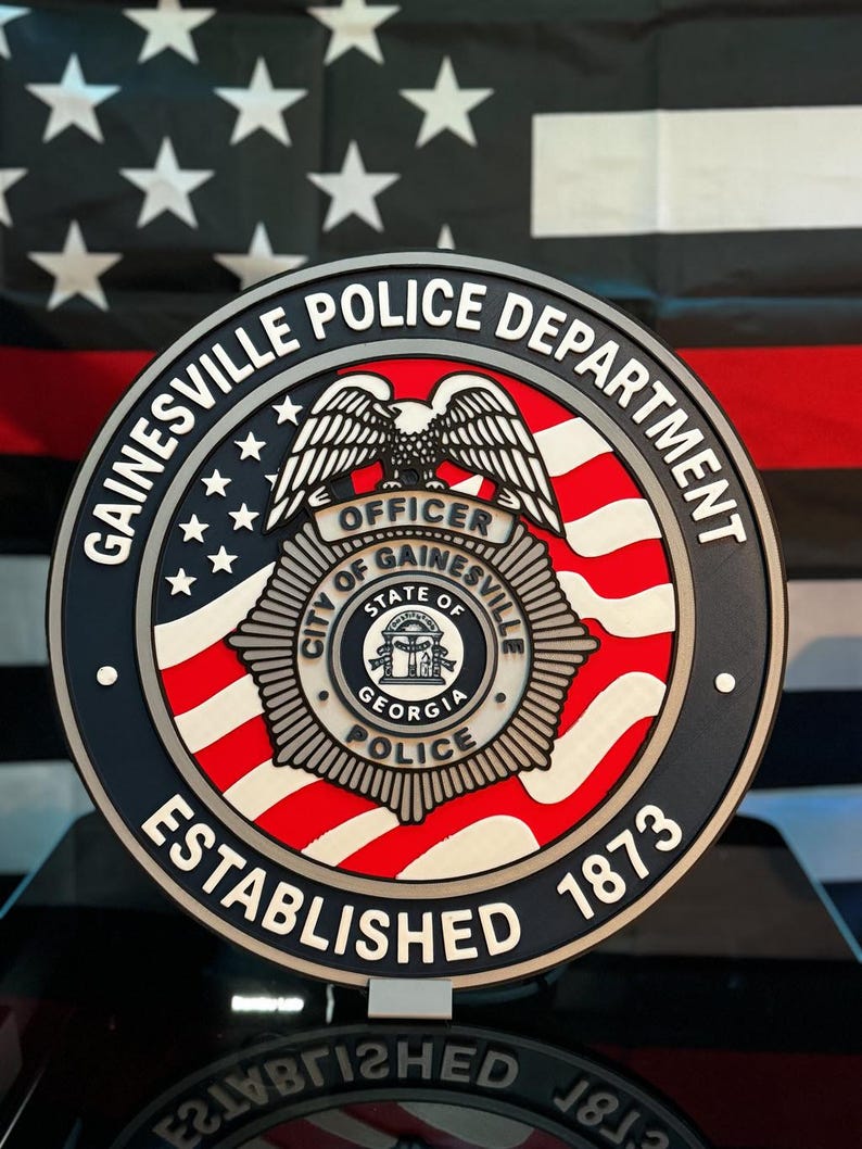 May include: A circular Gainesville Police Department emblem featuring a central badge with an eagle, the text City of Gainesville State of Georgia Police, and the words Gainesville Police Department Established 1873 on a red, white, and blue background.