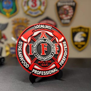 May include: A circular Streamwood Professional Firefighters Local 3022 emblem featuring a red and white Maltese cross design with black accents. The center displays the IAFF logo with the text International Association of Firefighters AFL-CIO CLC.