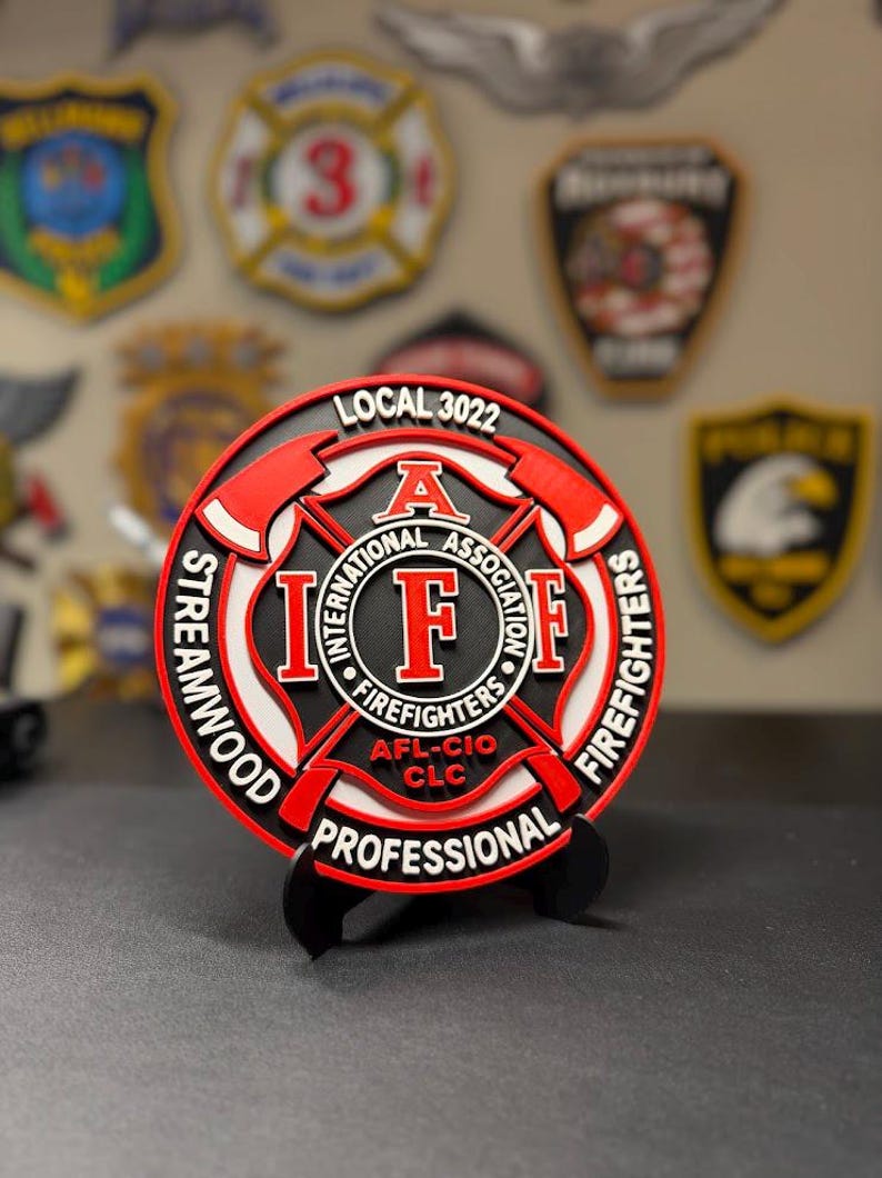 May include: A circular Streamwood Professional Firefighters Local 3022 emblem featuring a red and white Maltese cross design with black accents. The center displays the IAFF logo with the text International Association of Firefighters AFL-CIO CLC.