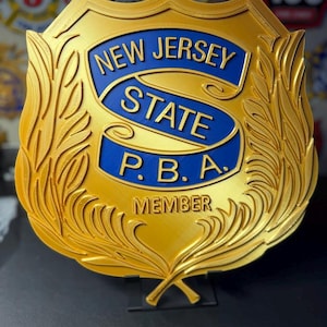 May include: A gold-colored shield-shaped emblem with the words "NEW JERSEY STATE P.B.A. MEMBER" in blue and gold. The emblem has a decorative leaf design around the edges.