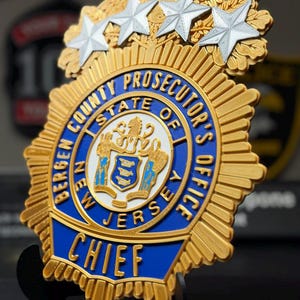 May include: A gold and blue Bergen County Prosecutor's Office Chief badge featuring four silver stars at the top. The central emblem displays the New Jersey state seal in white, blue, and gold, surrounded by text reading Bergen County Prosecutor's Office.