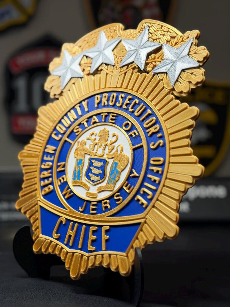 May include: A gold and blue Bergen County Prosecutor's Office Chief badge featuring four silver stars at the top. The central emblem displays the New Jersey state seal in white, blue, and gold, surrounded by text reading Bergen County Prosecutor's Office.