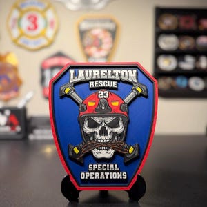May include: A shield-shaped plaque featuring a blue background with a red border. It displays a skull wearing a red helmet marked with the number 23, crossed axes, and the text LAURELTON RESCUE and SPECIAL OPERATIONS in white lettering.