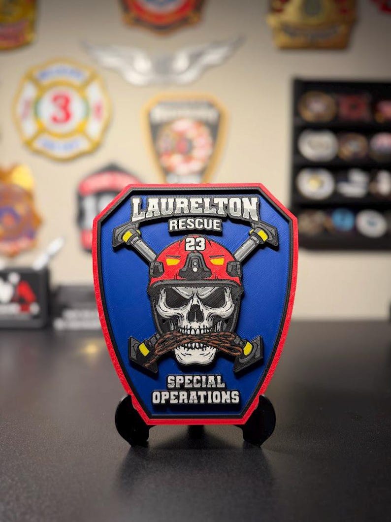 May include: A shield-shaped plaque featuring a blue background with a red border. It displays a skull wearing a red helmet marked with the number 23, crossed axes, and the text LAURELTON RESCUE and SPECIAL OPERATIONS in white lettering.
