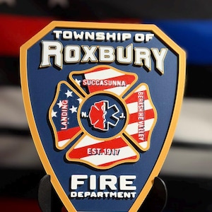May include: A shield-shaped emblem for the Township of Roxbury Fire Department with a gold border and blue background. The center features a Maltese cross with a red and white medical symbol, surrounded by text including Succasunna, Landing, and Berkshire Valley.