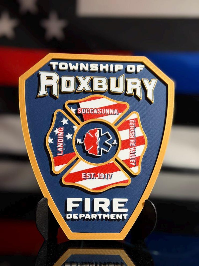 May include: A shield-shaped emblem for the Township of Roxbury Fire Department with a gold border and blue background. The center features a Maltese cross with a red and white medical symbol, surrounded by text including Succasunna, Landing, and Berkshire Valley.