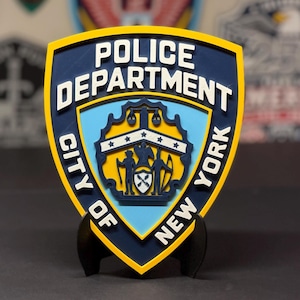 May include: A layered, shield-shaped sign featuring the New York City Police Department emblem. The design includes white raised lettering reading POLICE DEPARTMENT CITY OF NEW YORK on a dark blue background, with a central light blue crest.