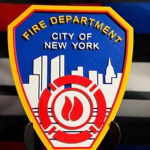 May include: A 3D-printed emblem for the New York City Fire Department. The shield-shaped design features the text "FIRE DEPARTMENT CITY OF NEW YORK" in yellow, a blue background, and a red and white fire symbol.