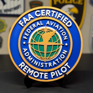 May include: A circular FAA Certified Federal Aviation Administration Remote Pilot sign featuring a blue background with gold and white lettering. The center displays a green globe graphic with a gold winged emblem and a crosshair overlay.