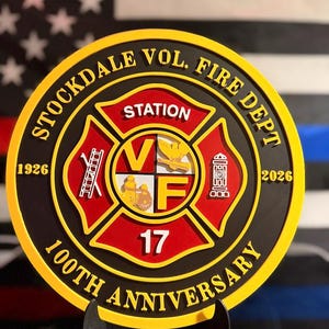 May include: A circular commemorative plaque for the Stockdale Vol. Fire Dept. 100th Anniversary, featuring a red and gold Maltese cross emblem with the text Station 17, a ladder, a fire hydrant, and the years 1926 and 2026 in raised yellow lettering.