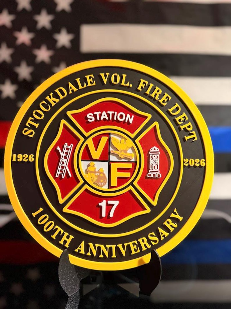 May include: A circular commemorative plaque for the Stockdale Vol. Fire Dept. 100th Anniversary, featuring a red and gold Maltese cross emblem with the text Station 17, a ladder, a fire hydrant, and the years 1926 and 2026 in raised yellow lettering.