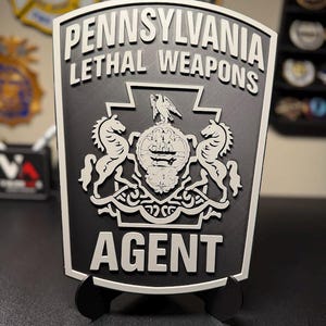 May include: A black and white shield-shaped plaque featuring the text PENNSYLVANIA LETHAL WEAPONS AGENT. The center displays the Pennsylvania state coat of arms with two horses, a central shield, and an eagle crest, all in a raised relief design.