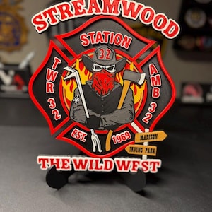 May include: A multi-layered wooden sign shaped like a Maltese cross featuring the text STREAMWOOD STATION, TWR 32, AMB 32, EST 1969, THE WILD WEST, MADISON, and IRVING PARK. The center depicts a figure in a cowboy hat, bandana, and skull mask.
