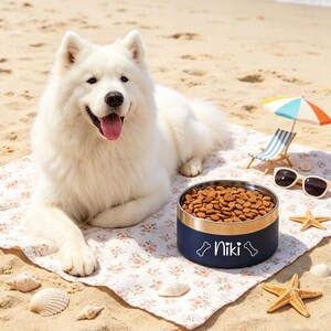 May include: A white Samoyed dog lies on a floral blanket on a sandy beach next to a navy blue dog bowl filled with kibble. The bowl has a gold rim and the name "Niki" printed on it. A miniature beach chair, sunglasses, and starfish are also present.