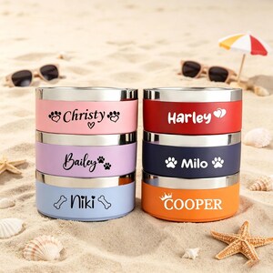 May include: A stack of six personalized pet bowls in various colors, including pink, red, blue, and orange. Each bowl has a silver rim and features a pet's name with paw prints or a bone graphic. The bowls are displayed on a sandy surface with sunglasses and a small umbrella.