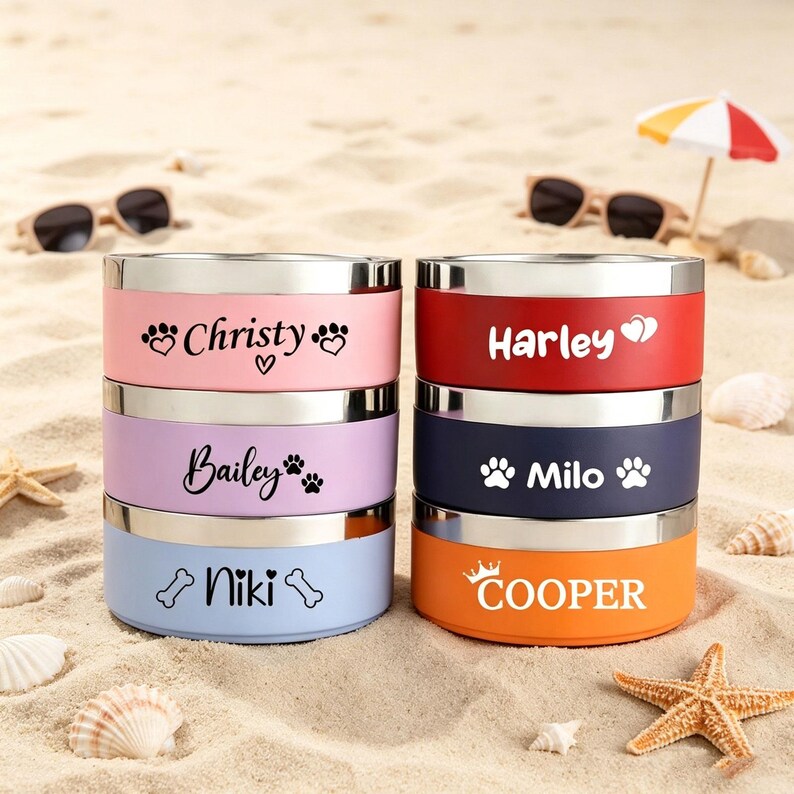 May include: A stack of six personalized pet bowls in various colors, including pink, red, blue, and orange. Each bowl has a silver rim and features a pet's name with paw prints or a bone graphic. The bowls are displayed on a sandy surface with sunglasses and a small umbrella.