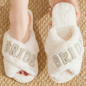 May include: White fluffy cross-band slippers with the word "BRIDE" spelled out in sparkling rhinestones. The slippers are made of a soft, plush material and are designed for comfort. Perfect for a bridal shower or wedding day.