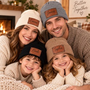 May include: Four ribbed knit beanies in cream, gray, black, and tan, each featuring a rectangular brown leather patch with a name engraved in black script. The names Emma, James, Noah, and Sophie are clearly visible on the respective patches.