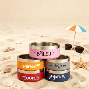 May include: A stack of stainless steel pet bowls in various colors, including pink, orange, gray, red, and navy blue. Each bowl has a name printed on it: Bailey, Harley, Christy, Cooper, and Julie. The bowls are on a sandy surface.