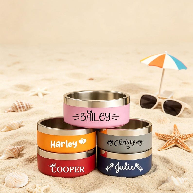 May include: A stack of stainless steel pet bowls in various colors, including pink, orange, gray, red, and navy blue. Each bowl has a name printed on it: Bailey, Harley, Christy, Cooper, and Julie. The bowls are on a sandy surface.