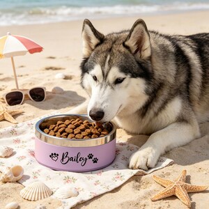 May include: A lilac pet bowl with a silver rim, filled with dog food, sits on a beach. The bowl is personalized with the name "Bailey", a heart, and paw prints. A gray and white husky dog is eating from the bowl. Sunglasses and a small beach umbrella are in the background.