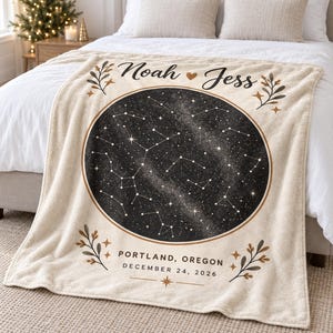 Könnte beinhalten: Eine cremefarbene Fleecedecke mit einer zentralen, kreisförmigen schwarzen Sternenkarte. Der Text lautet Noah und Jess mit einem Herz, Portland, Oregon, und 24. Dezember 2026. Dekorative Zweige und kleine Sterne umrahmen das Design.