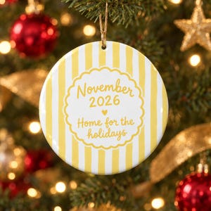 Può includere: Una decorazione natalizia rotonda in ceramica con strisce verticali gialle e bianche. Il centro presenta una cornice smerlata gialla con il testo November 2026, un piccolo cuore e la frase Home for the holidays in corsivo giallo.