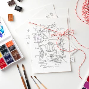 May include: A black and white line drawing of a window with a cactus in a teacup, a birdhouse, and a small plant. The drawing is on a white sheet of paper with red and white twine tied around it. There are paintbrushes and watercolor paints on the table.