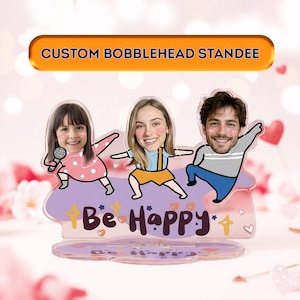 Puede incluir: Un expositor de bobblehead personalizado con tres caras sonrientes. El expositor tiene una base morada con las palabras "Be Happy" y un diseño decorativo. El banner superior dice "CUSTOM BOBBLEHEAD STANDEE".
