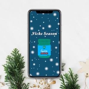 May include: A smartphone displays a teal background with white snowflakes and the text "Vicks Season." The image features a green jar of VapoRub with a blue label. The phone is surrounded by Christmas decorations.
