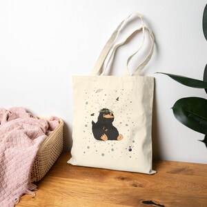May include: A cream-coloured tote bag with a charming illustration of a black creature wearing a floral crown. The design includes small birds and scattered dots. The bag has long handles and is made of a sturdy fabric, ideal for carrying items.