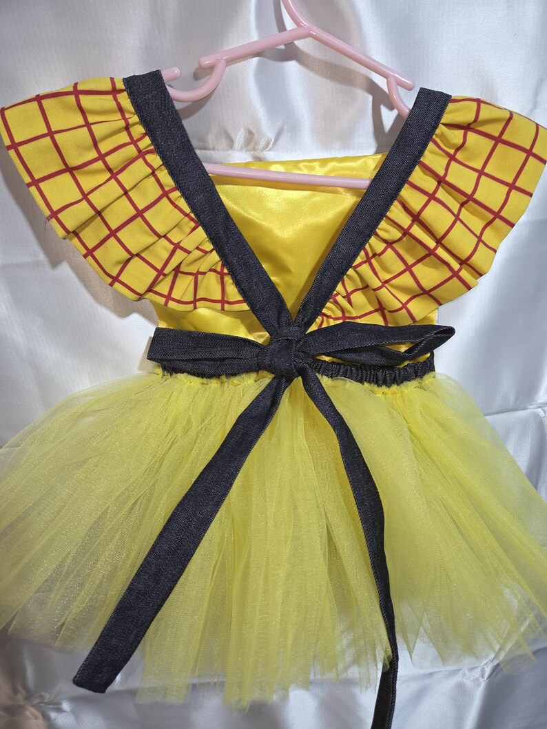 May include: A yellow and red plaid dress with a tulle skirt, reminiscent of a cartoon character. The dress features a dark gray bow and straps, and is hanging on a pink hanger.