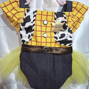 May include: A baby-sized costume resembling a sheriff's outfit, featuring a yellow plaid shirt with red lines, a cow-print vest, and a denim bottom. The costume includes a brown belt with a decorative buckle, a gold sheriff badge, and a yellow tulle skirt.