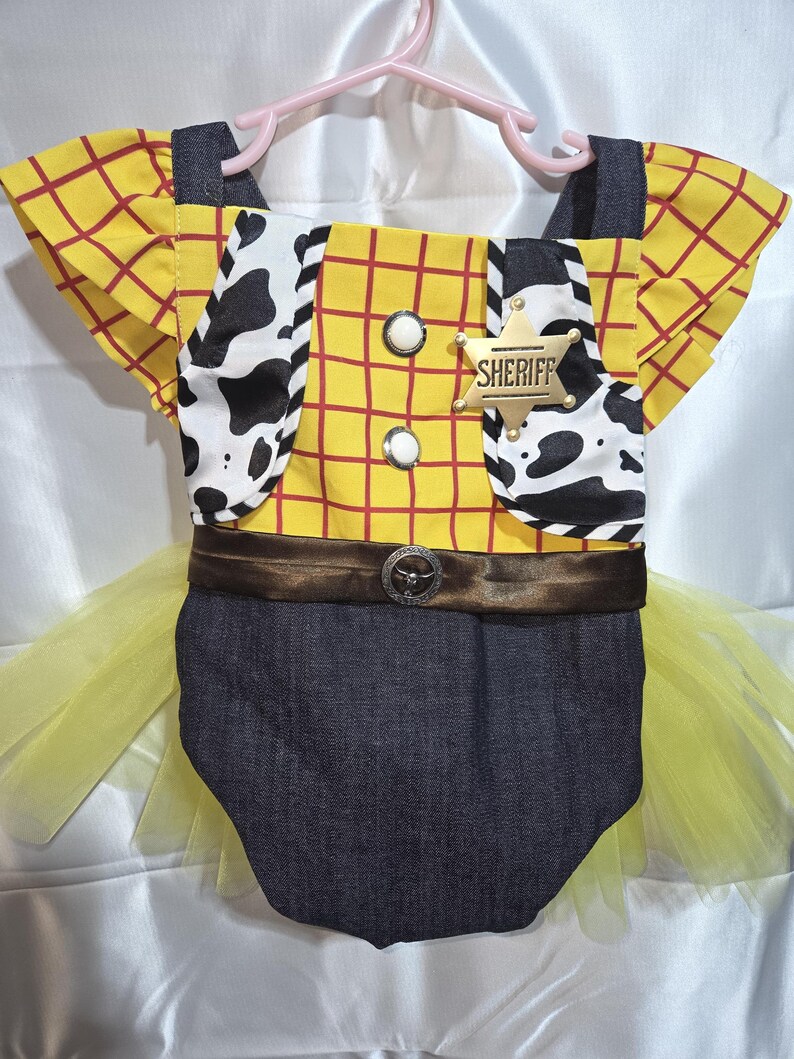 May include: A baby-sized costume resembling a sheriff's outfit, featuring a yellow plaid shirt with red lines, a cow-print vest, and a denim bottom. The costume includes a brown belt with a decorative buckle, a gold sheriff badge, and a yellow tulle skirt.