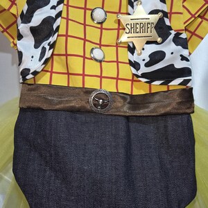 May include: A yellow and red plaid bodysuit with a brown belt and a Sheriff badge. The bodysuit has a cow print vest and a denim bottom. The Sheriff badge is gold with the word "Sheriff" on it. The bodysuit is a costume.