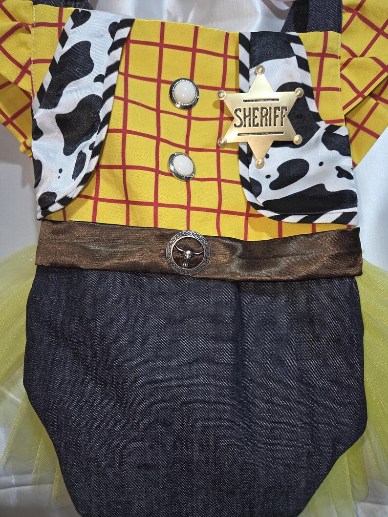 May include: A yellow and red plaid bodysuit with a brown belt and a Sheriff badge. The bodysuit has a cow print vest and a denim bottom. The Sheriff badge is gold with the word "Sheriff" on it. The bodysuit is a costume.
