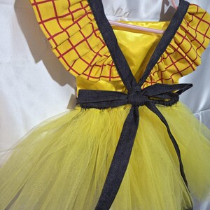 May include: A yellow and red plaid dress with a black bow and straps. The dress has a yellow tulle skirt and is hanging on a pink hanger. The top of the dress has a yellow background with a red plaid pattern.