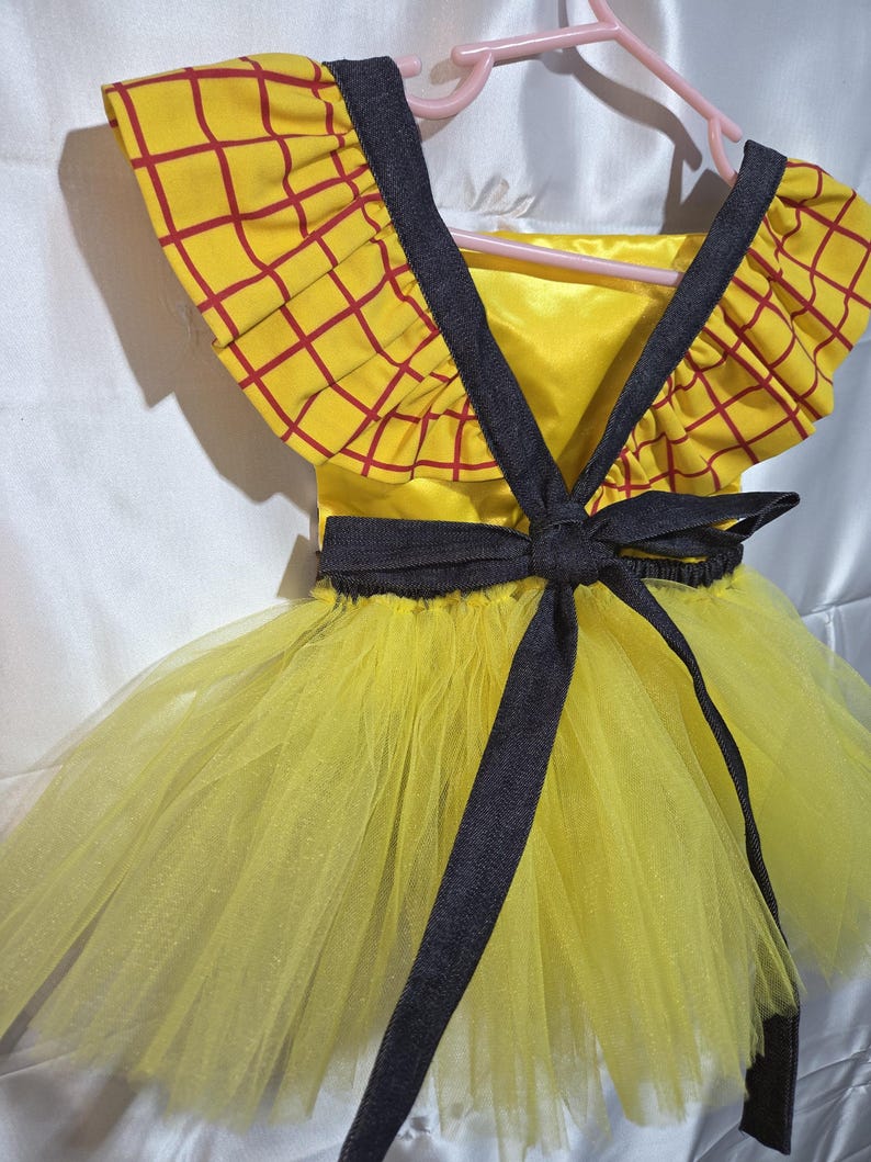 May include: A yellow and red plaid dress with a black bow and straps. The dress has a yellow tulle skirt and is hanging on a pink hanger. The top of the dress has a yellow background with a red plaid pattern.