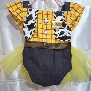May include: A yellow and red plaid romper with a Sheriff badge, cow print accents, and a denim bottom. The outfit includes a brown belt and a yellow tulle skirt, hanging on a pink hanger.