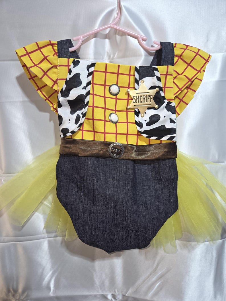 May include: A yellow and red plaid romper with a Sheriff badge, cow print accents, and a denim bottom. The outfit includes a brown belt and a yellow tulle skirt, hanging on a pink hanger.